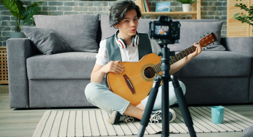 A young musician sits on the floor, playing guitar and recording a video