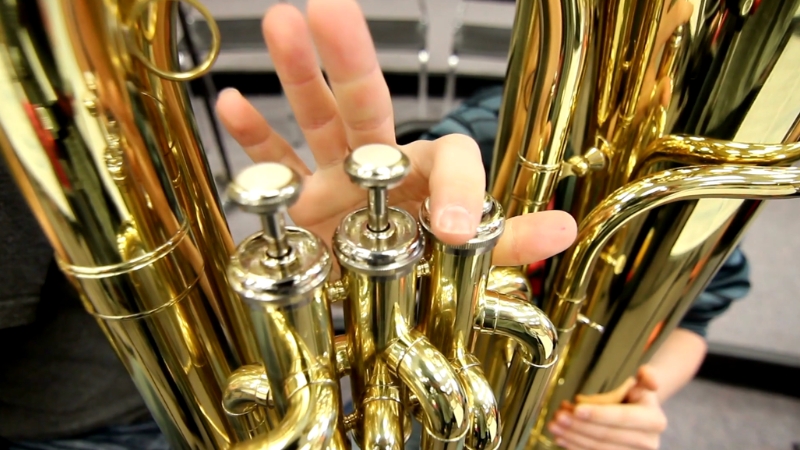 A close-up shows a person pressing the valves of a brass instrument during practice