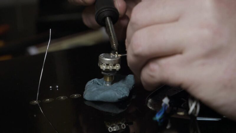 Close-up of hands soldering wires to a potentiometer on a guitar body
