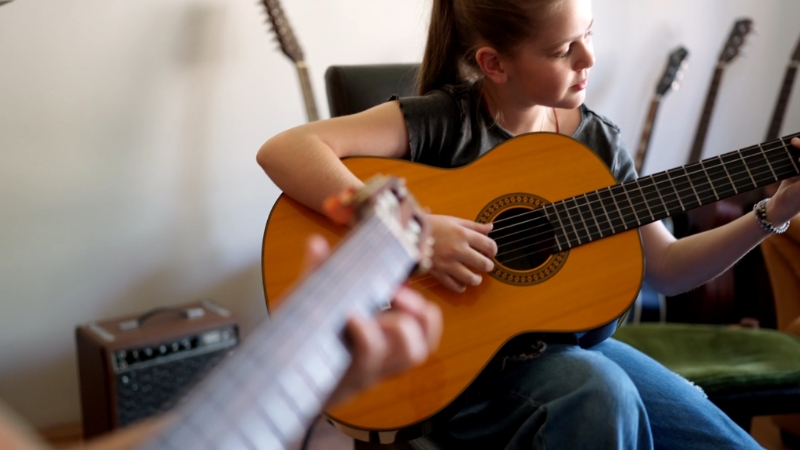 A young girl practices guitar during a lesson, with another player guiding her from the side