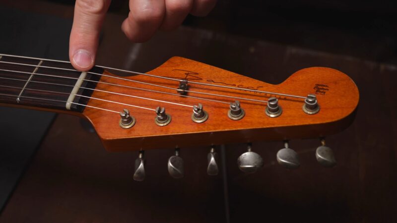 Close-up of a hand pointing at the nut on a guitar headstock with tuning pegs and strings visible.