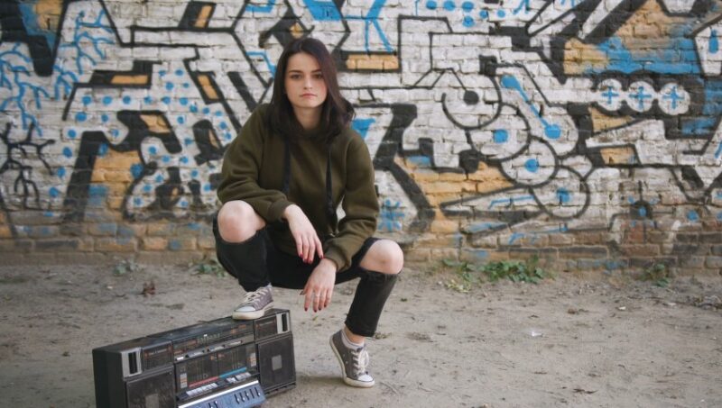 Young woman sits atop a vintage boombox, against a vibrant graffiti-covered wall