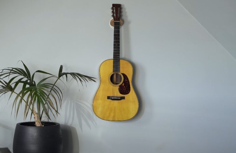 A light-toned acoustic guitar hangs on a light gray wall