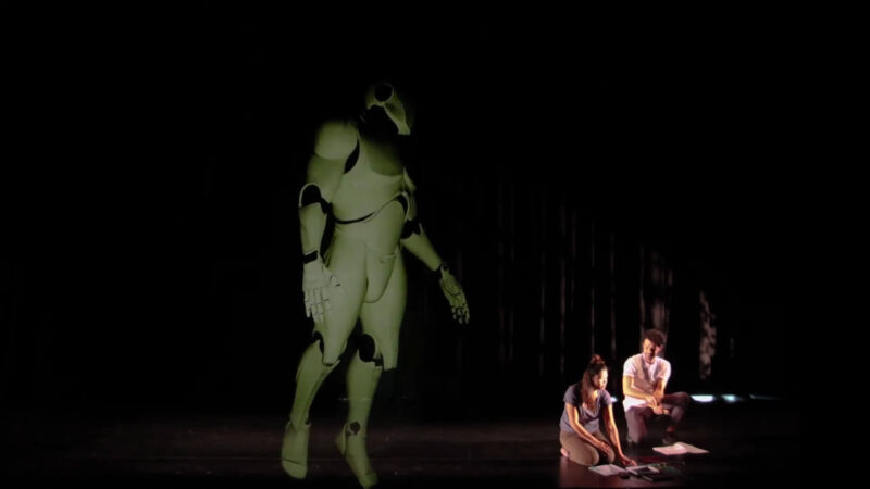 Two figures sit on the stage floor, observing a large, articulated robot figure