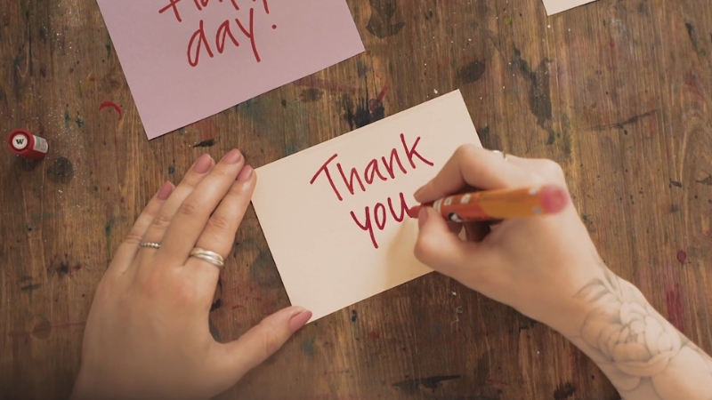 A person writes โThank youโ in red marker on a card at a wooden table with other notes nearby