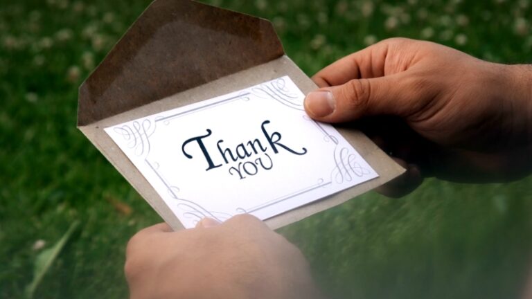 Hands hold an open envelope with a thank you card inside against a grassy background