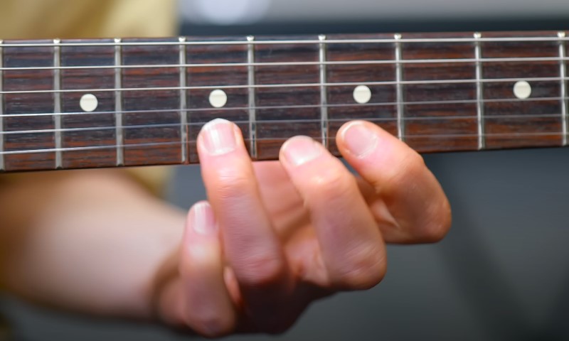 Close-up of fingers pressing down on a dark wooden guitar fretboard near the fifth fret marker