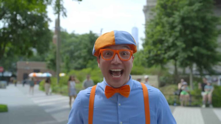 Person wearing an orange-and-blue hat, orange glasses, and bow tie, smiling energetically outdoors in a park setting