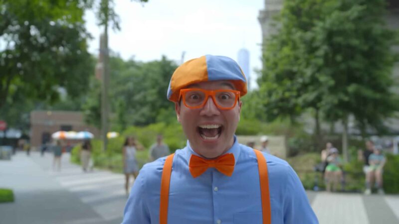 Person wearing an orange-and-blue hat, orange glasses, and bow tie, smiling energetically outdoors in a park setting