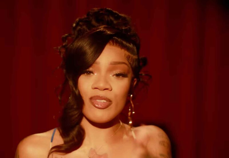 A woman performing in front of a red background, confidently facing the camera with styled hair and makeup