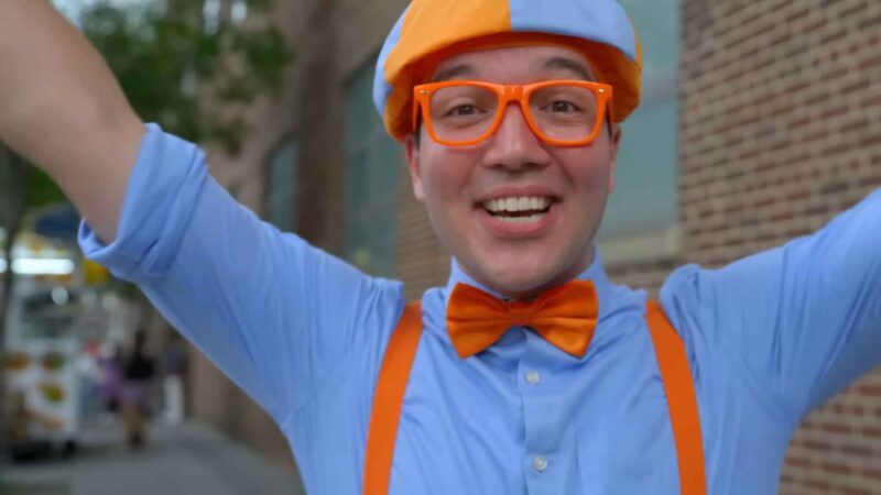 Person wearing a blue and orange outfit with matching hat, glasses, bow tie, and suspenders, smiling outdoors with arms raised in excitement