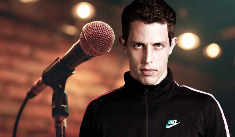 Man in a black Nike jacket standing in front of a microphone on stage with blurred lights in the background