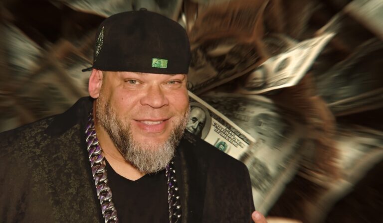 Man wearing a black hat and chain smiling in front of a blurred background of U.S. dollar bills