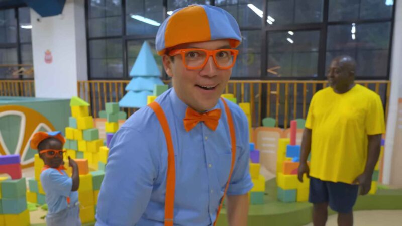 Person dressed in a blue and orange outfit with a bow tie, suspenders, and hat, standing in a colorful play area with children and adults in the background