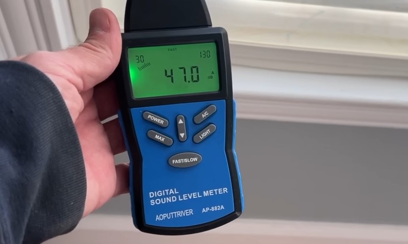 A person holds a digital meter in front of a window, measuring sound