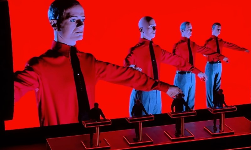 A red screen displays three men in suits standing on stage, representing the electronic music group Kraftwerk