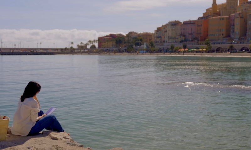 Woman reading by the shimmering turquoise water with the colorful town of Menton rising in the background