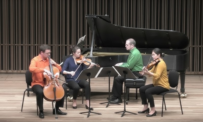 Four musicians playing violin, cello, and piano in front of a grand piano, creating a harmonious ensemble performance