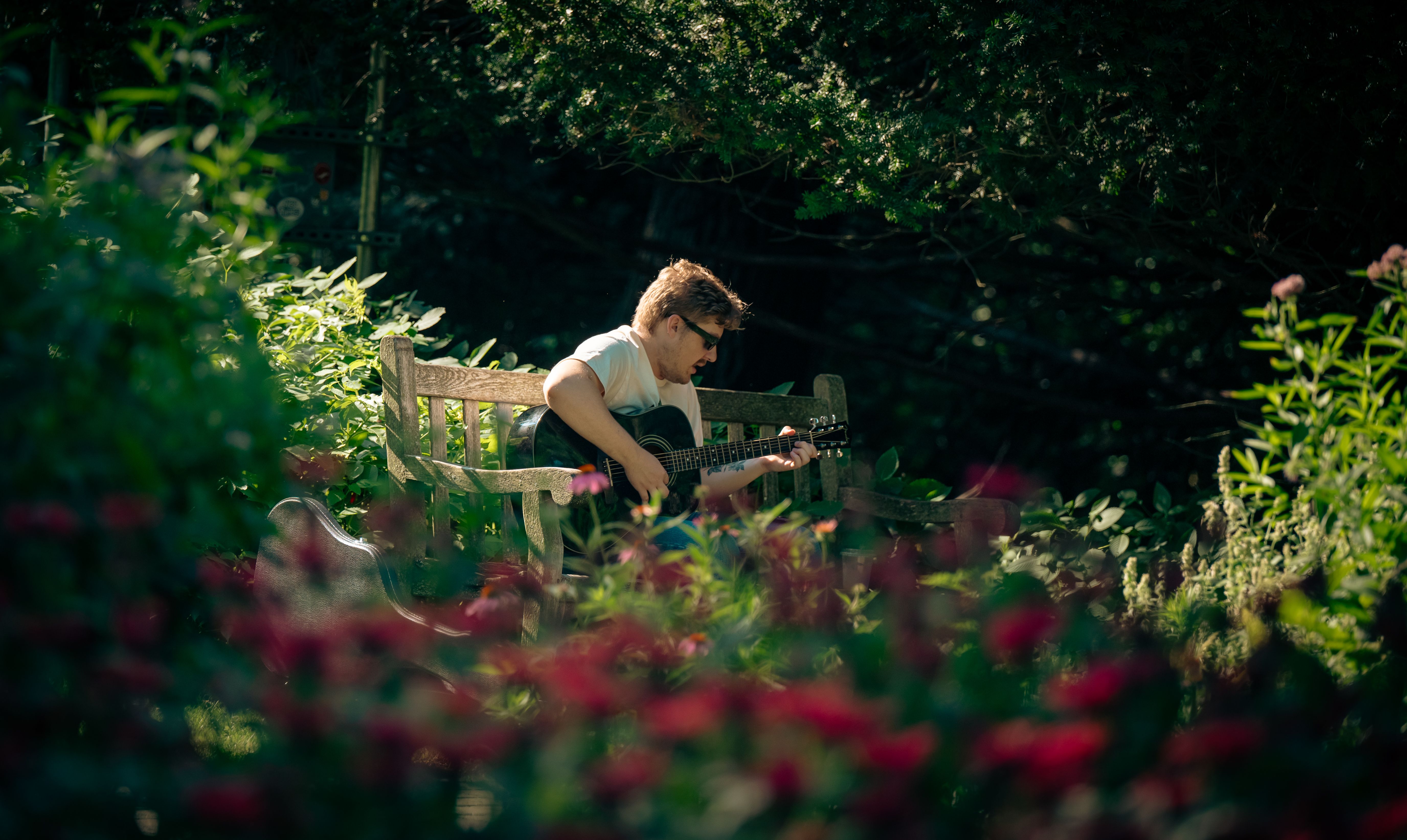A man plays acoustic guitar on a garden bench surrounded by greenery